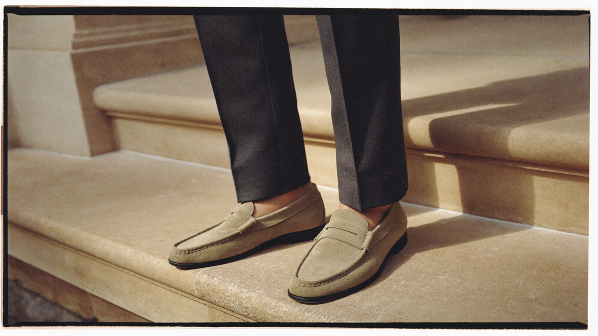 Sand suede loafers worn without socks with charcoal tailored trousers on a stone step