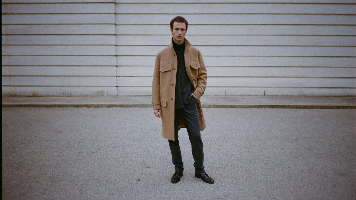 A man in a plain camel overcoat and charcoal trousers on an empty pavement, no visible branding