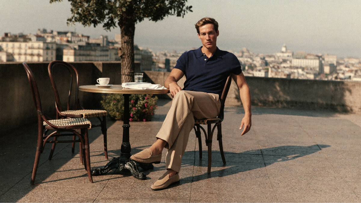 A man in a navy pique polo shirt and sand tailored trousers seated at an outdoor cafe terrace